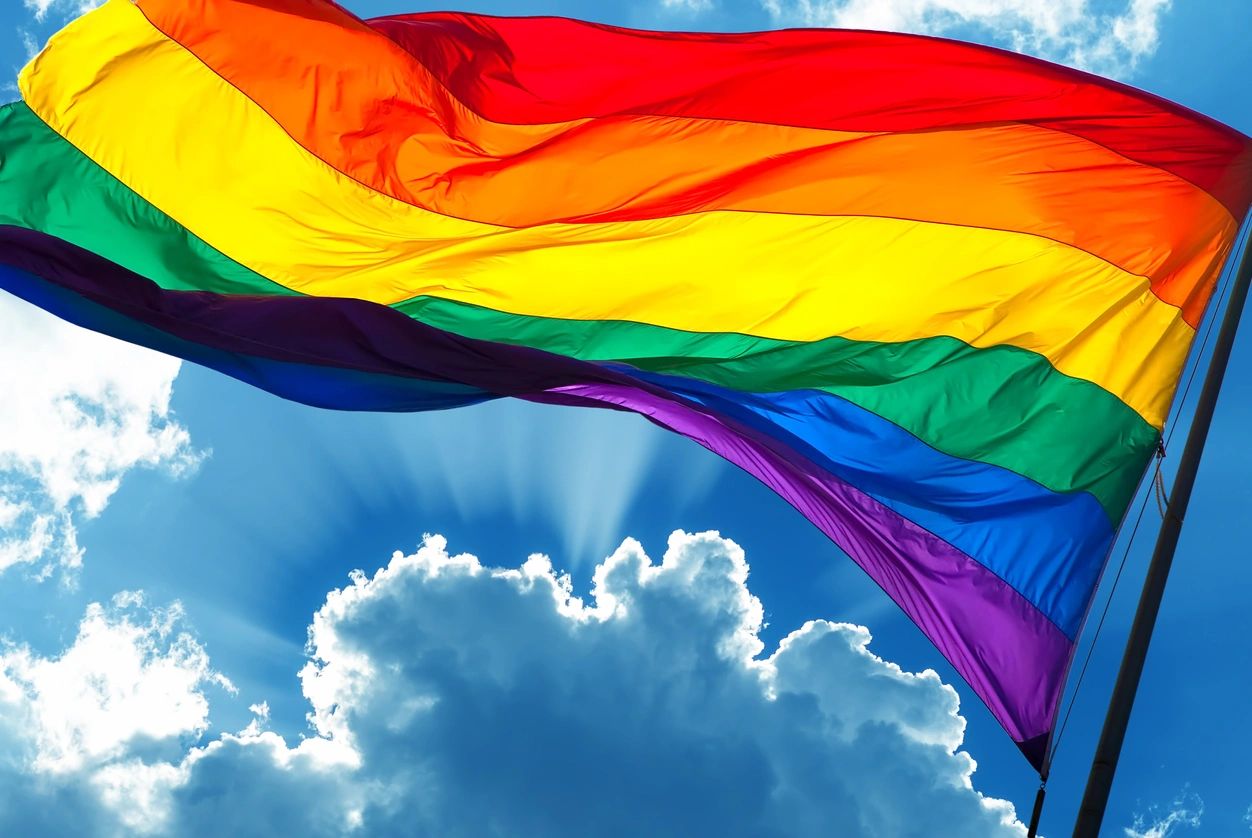 A rainbow flag flying in the sky with clouds behind it.