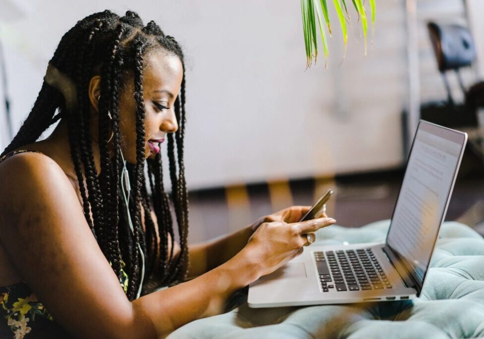 A woman is looking at her phone while using a laptop.